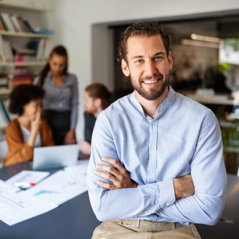 Mann sitzt mit verschränkten Armen am Tisch, im Hintergrund Kollegen im Meeting